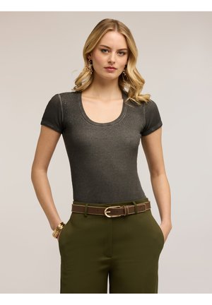 Blondýna nosící přiléhavé tmavě šedé tričko s krátkými rukávy zdobené malými korálky, olivově zelené kalhoty, hnědý opasek a zlaté šperky.