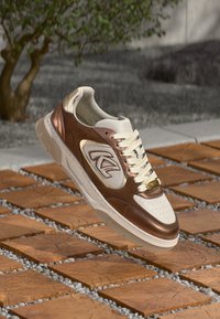 Αθλητικό παπούτσι με λευκό και καφέ δερμάτινο επάνω μέρος, διάτρητο κουτί δαχτύλων, ανασηκωμένο λογότυπο, μπεζ κορδόνια και χρυσή λεπτομέρεια στη σόλα.