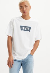 Balta medvilninė marškinė su mėlynu Levi's logotipu ant krūtinės. Marškiniai turi trumpas rankoves ir apvalų iškirpimą. Dėvimi su šviesiai mėlynomis džinsais.