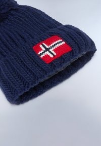 Berretto in maglia blu navy con una texture a coste e un patch rosso con un motivo a croce bianco. Materiale morbido, forma arrotondata e visiera ripiegata.