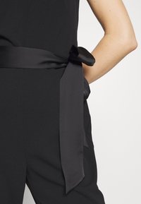 Ceinture en tissu noir nouée en un nœud autour de la taille d'une personne portant des vêtements noirs, un bras reposant sur la hanche.
