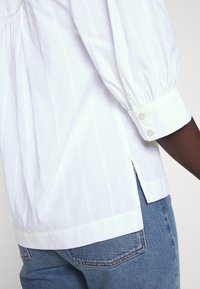 Hvitstripet bluse med puffermer, tettsittende mansjetter med to knapper, og sidesplits. Kombineres med blå denimjeans som er synlige nederst.