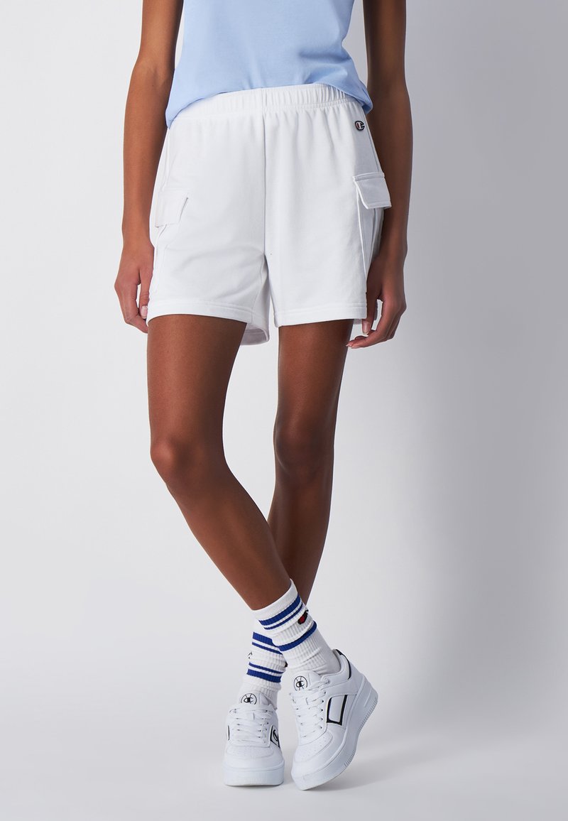 Pantaloni corti bianchi in cotone con una cintura elastica, dotati di due tasche laterali. Abbinati a sneaker bianche e calzini a righe.