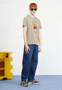 Beige graphic t-shirt met een NASA ontwerp en hemelse afbeeldingen, gecombineerd met losse blauwe spijkerbroeken en multicolore sneakers. Donkere zonnebril op.