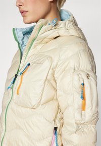 Lichtbeige pufferjack met capuchon, voorzien van meerdere ritszakken, kleurrijke ritsen in blauw, oranje en groen, en een gestikte structuur.