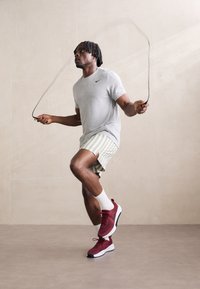 Sporty røde og hvide sko, grå T-shirt og shorts med grønne striber. Personen hopper sejr i sjippetov mod en ensfarvet baggrund.