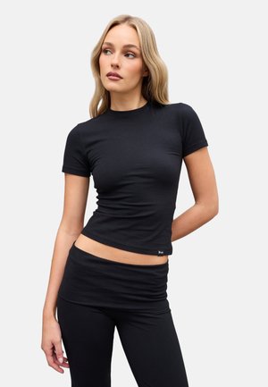 Mujer con cabello rubio largo que lleva una camiseta negra ajustada de manga corta y pantalones negros a juego sobre un fondo liso.