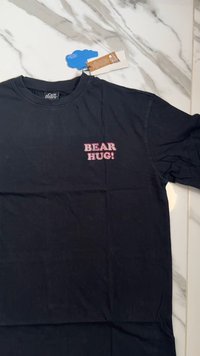 Must musta t-särk mustast puuvillast, mille esiosas on roosa trükitud graafika, millel on kiri "BEAR HUG!" Lühikesed varrukad, ümmargune kaelus ja sile tekstuur.