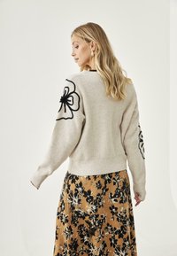 Suéter de punto color beige claro con textura acanalada, decorado con bordados florales negros en las mangas; combinado con una falda de estampado floral.