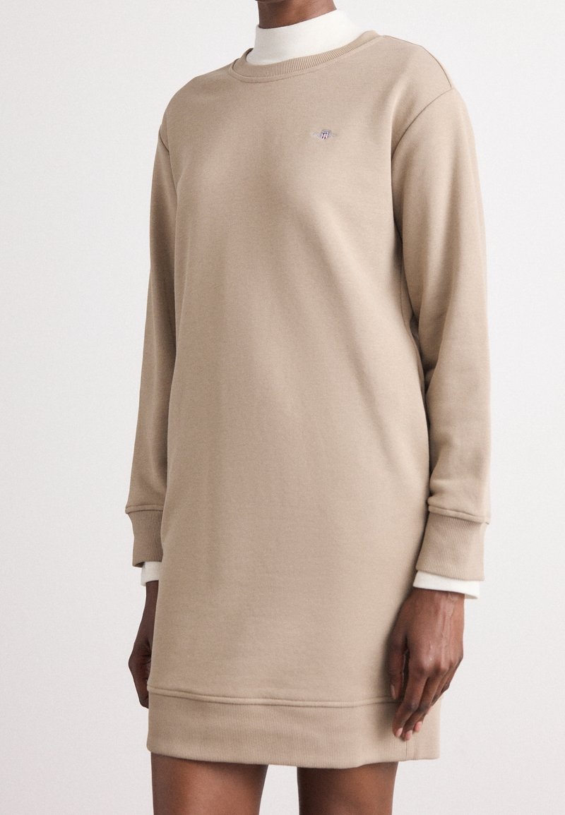 Robe sweat beige à manches longues portée sur un col roulé blanc, avec poignets côtelés et petit logo sur la poitrine.