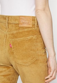 Gros plan d'une personne portant un pantalon beige en velours côtelé Levi's avec une poche arrière visible, un patch de la marque et un haut blanc côtelé sans manches.