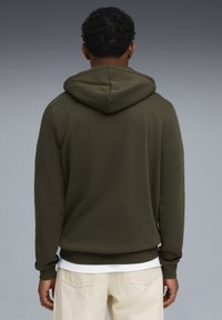 Felpa con cappuccio in verde oliva scuro, realizzata in morbido tessuto. Presenta una tasca a canguro e polsini a costine. Abbinata a strati sotto una t-shirt bianca.