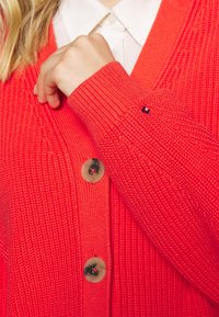 Cárdigan rojo brillante con tejido texturizado, que presenta un escote en V profundo y un cierre de grandes botones. Incluye un pequeño acento de logo en la manga.