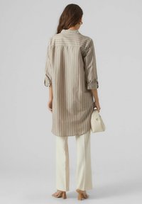 Woman wearing a beige striped long shirt, white pants, beige heels, holding a cream handbag, standing against a plain light grey background.