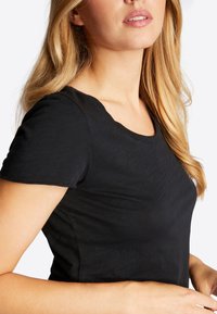 Lyshåret kvinde iført en ensfarvet sort t-shirt med korte ærmer, med let åben mund, beskåret fra skuldrene til midten af overkroppen.