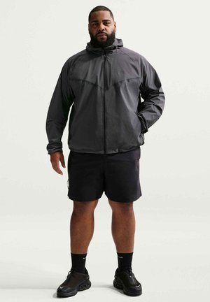 Man met baard die een zwarte hoodie, zwarte korte broek, zwarte sokken en zwarte sportschoenen draagt, staand tegen een effen achtergrond.