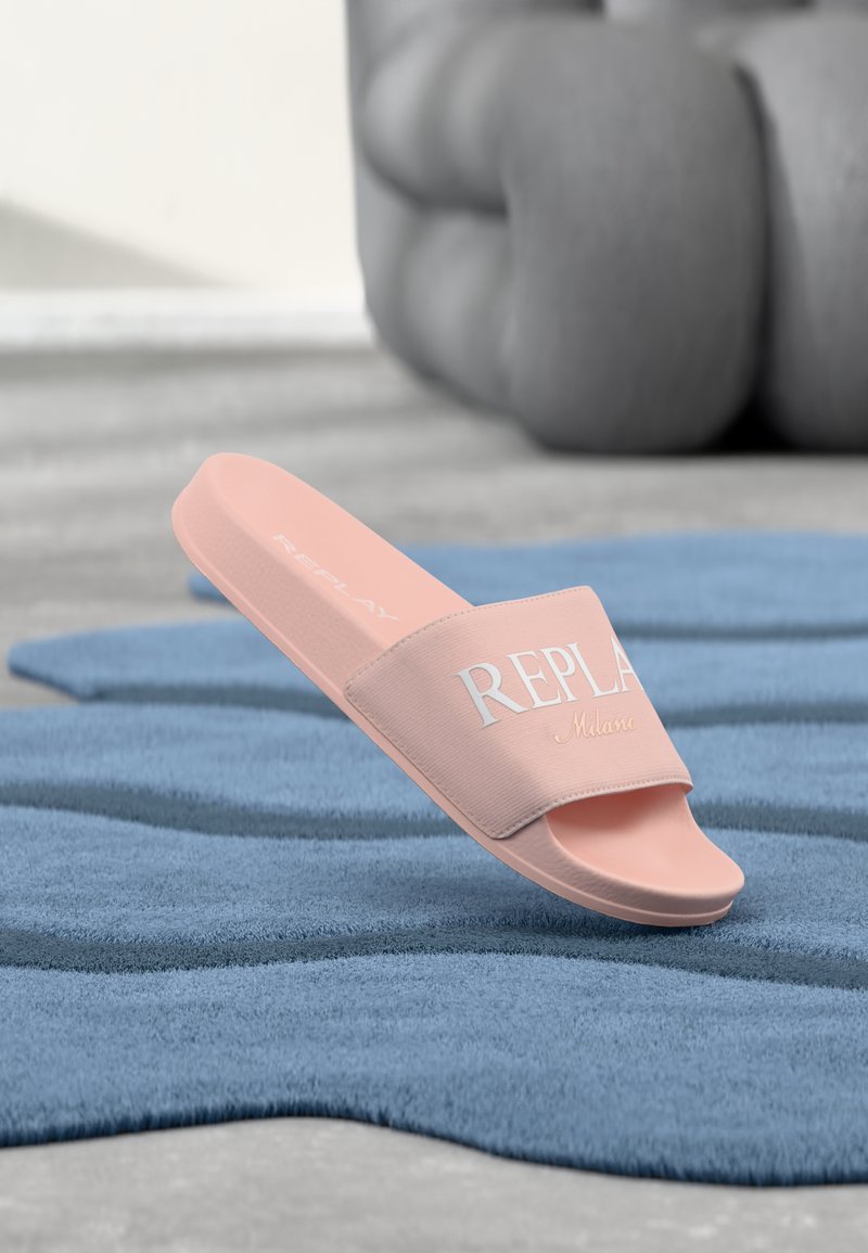 Μονό ροζ σαγιονάρι με το κείμενο "REPLAY Milano" στο λουράκι, ακουμπισμένο σε υφή μπλε επιφάνειας με θολό φόντο.