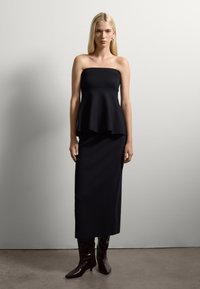 Una donna che indossa un top peplum nero senza spalline, una lunga gonna nera e stivali neri a punta, in piedi contro un muro bianco semplice.