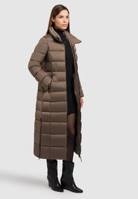 Ilgas rudas pūkinis paltas su aukštu apykakle, vertikaliu siūlomu raštu, šoninėmis kišenėmis ir užtrauktukais. Dėvimas virš juodos megztos suknelės ir juodų batus.