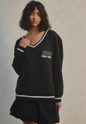 Svart oversize sweatshirt med V-hals, vit kant och grafiskt tryck "1990 NEW YORK". Bärs med en svart, rynkad kjol. Mjuk tygtextur.