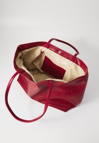 Sac fourre-tout en cuir tressé rouge avec deux poignées robustes. L'intérieur est doté d'une doublure beige et d'une poche zippée avec un accent rouge.
