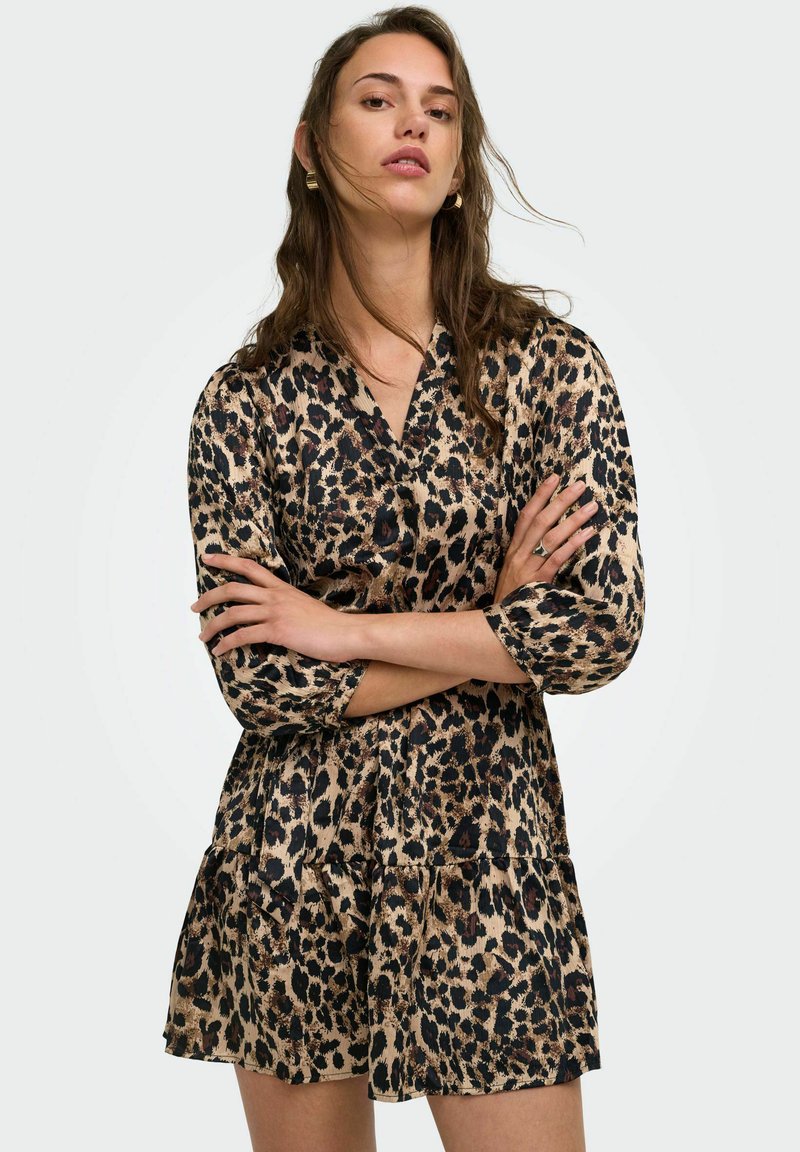 Jeune femme aux longs cheveux portant des boucles d'oreilles créoles dorées et une robe à imprimé léopard avec des manches trois-quarts, bras croisés, fond neutre.