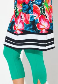 Kwiatowa tunika w czerwone, różowe i niebieskie kwiaty na czarnym tle, zestawiona z zielonymi legginsami 3/4 z rozcięciem na dole.