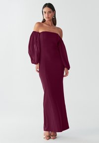 Bordo brez naramnic obleka z oprijetim bodijem, volumniziranimi prosojnimi nabranimi rokavi in ravno krilo do tal. Oblečena z metalik petami.