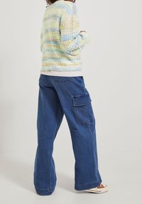 Mnohobarevný pletený svetr s žlutými, modrými a zelenými pruhy; spojený s širokými džíny z modrého denimu s kapsami po stranách.