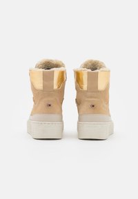 Un paio di stivaletti beige in suede con accenti metallici oro, suole spesse bianche e interni foderati in pile, mostrati da dietro su uno sfondo bianco.