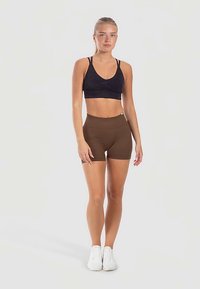 Schwarzer Sport-BH mit V-Ausschnitt und verstellbaren Trägern, kombiniert mit braunen High-Waist-Shorts. Das Modell trägt weiße Sportschuhe.