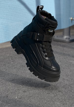 Black high-top boot featuring a textured leather upper, chunky sole, lace and strap closure, with a pull tab at the heel and accent stitching.