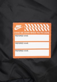Nike-märke på svart tyg med orange kant, vit text "LÄMNA ÖVER NÄR DU ÄR KLAR," och tre tomma fält för "FÖREDRAGET NAMN:".