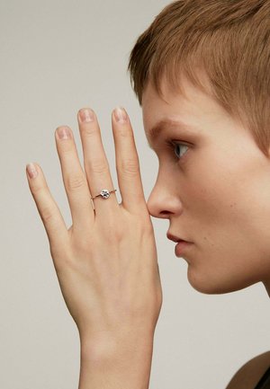 Anillo de plata con una piedra preciosa central, llevado en un dedo. La mano es esbelta, con uñas cortas. El fondo es neutro.