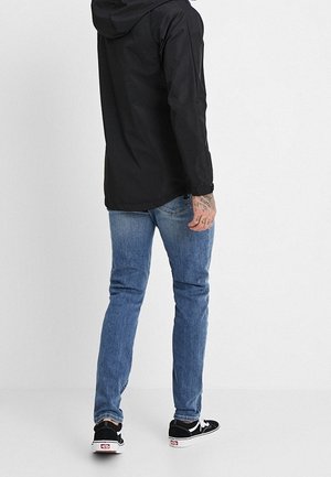 Homme portant une veste à capuche noire, un jean bleu et des baskets noires avec des semelles blanches, debout de dos sur un fond clair uni.