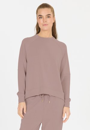 Vrouw met achtergebonden haar die een mauve longsleeve sweater en bijpassende broek met trekkoord draagt tegen een effen witte achtergrond.