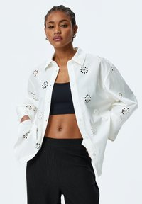 White button-up shirt with floral cut-out details, featuring a relaxed fit and long sleeves, worn over a black ribbed top and pants.