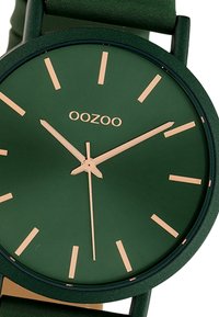 Montre verte avec finition mate, cadran rond, accents dorés et marqueurs d'heure simples. Elle dispose d'un bracelet en tissu et d'éléments de design minimalistes.