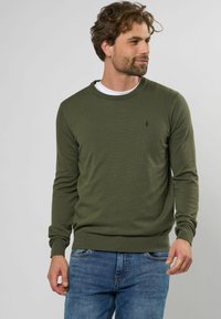 Suéter de punto en verde oliva con cuello y puños acanalados, que presenta un pequeño logo negro en el pecho. Lleva puesto sobre una camisa blanca, combinado con jeans azules.