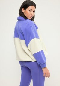 Lila und cremefarbener Fleece-Pullover mit hohem Kragen, Farbblock-Design und elastischen Bündchen. Kombiniert mit passenden lila Leggings.