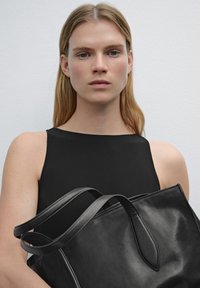 Sac en cuir noir avec deux longues poignées, texture lisse et coutures discrètes, tenu par une personne portant un haut sans manches noir.