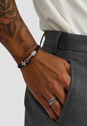 Sort flettet armbånd med sølvbeslag, der har en dekorativ lukning og knude. Hånd, der viser en sølvring med dobbelt bånd. Grå bukser.