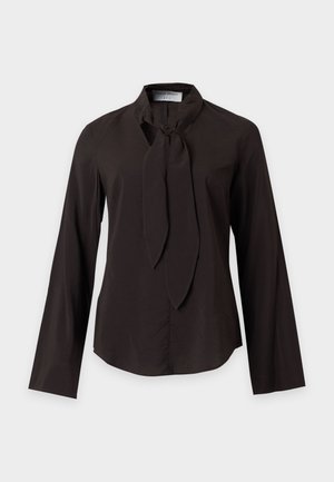 Donna con i capelli corti in piedi che indossa una maglia nera a maniche lunghe con scollo annodato, jeans larghi azzurri chiari e scarpe nere con punta appuntita.