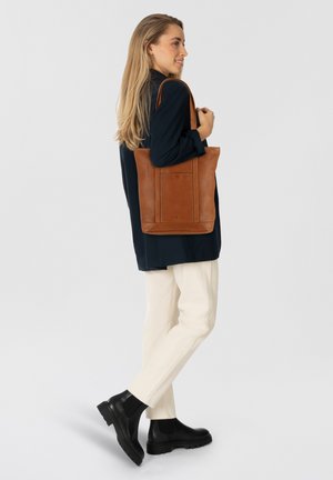 Bruine leren tote bag met verticale naden, een platte basis en stevige stoffen handvatten, gecombineerd met crèmekleurige broeken en zwarte enkellaarsjes.