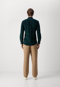 Camisa xadrez de manga longa verde escura com colarinho abotoado, usada com calças bege. Apresenta punhos com botão e um padrão de xadrez subtil.