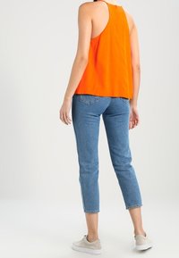 Top arancione senza maniche con una vestibilità rilassata, abbinato a jeans blu a vita media, con taglio dritto e lunghezza alla caviglia, completato da sneakers beige.
