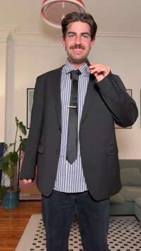 Čierne sako cez modro-bielu prúžkovanú košeľu s gombíkmi, so čiernou kravatou zdobenou striebornou sponou na kravatu. Tmavé džínsy, vnútorné prostredie.