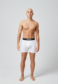 Boxer in bianco con elastico nero con la scritta "SNOCKS". Il tessuto sembra liscio, aderente e elasticizzato, con un taglio a metà coscia.