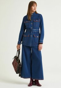 Giacca di jeans con quattro tasche e cintura, colore blu con cuciture rosse a contrasto; abbinata a pantaloni ampi in denim e stivali color bordeaux.