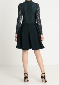 Mörkgrön klänning med ett spetsöverdel och långa ärmar, med en utsvängd kjol och veckdetaljer. Avslutad med en dragkedja bak.